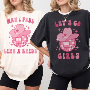 K&ouml;nnte beinhalten: Zwei Frauen tragen &uuml;bergro&szlig;e T-Shirts mit unterschiedlichen Designs. Das Shirt auf der linken Seite ist elfenbeinfarben und tr&auml;gt den Schriftzug "Man I feel like a bride" mit einer rosa Discokugel und einem Cowboyhut. Das Shirt auf der rechten Seite ist schwarz und tr&auml;gt den Schriftzug "Let's go girls" mit einer rosa Discokugel und einem Cowboyhut.