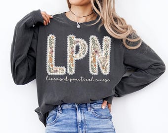 May include: Dark gray long-sleeve sweatshirt with the letters "LPN" in a floral design. The letters are outlined with a white lace trim. Below the letters is the text "licensed practical nurse" in a script font.