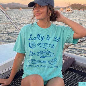 May include: A turquoise t-shirt with the text "Salty & 30" and fish illustrations. The shirt also includes the phrase "packed with flavour since 1996". The shirt is a comfortable, casual style.