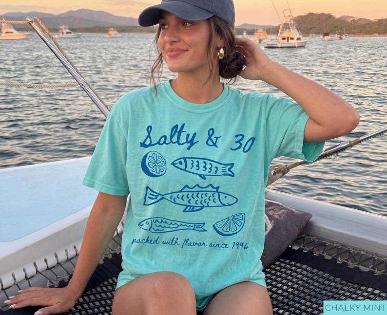 May include: A turquoise t-shirt with the text "Salty & 30" and fish illustrations. The shirt also includes the phrase "packed with flavour since 1996". The shirt is a comfortable, casual style.