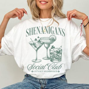 May include: White t-shirt with a green graphic design that says "Shenanigans Social Club" and features three cocktail glasses with shamrock accents. The text below the glasses says "Let's Get Shamrocked."