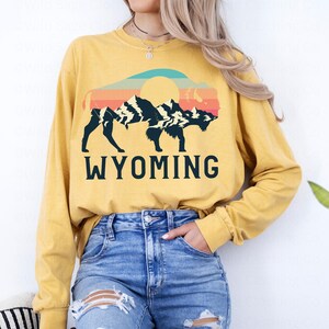May include: A yellow long-sleeved t-shirt with a vintage-style graphic of a bison silhouette in front of a mountain range and sunset. The text "WYOMING" is printed below the graphic.