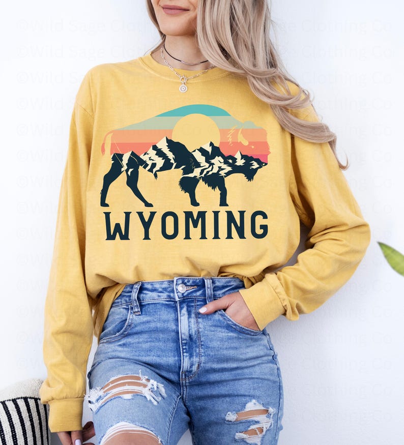 May include: A yellow long-sleeved t-shirt with a vintage-style graphic of a bison silhouette in front of a mountain range and sunset. The text "WYOMING" is printed below the graphic.