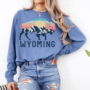 May include: A blue long-sleeved shirt with a graphic of a silhouette of a bison with a sunset and mountain range inside. The text "WYOMING" is printed below the graphic.