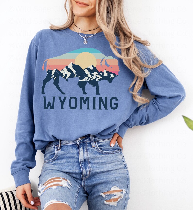 May include: A blue long-sleeved shirt with a graphic of a silhouette of a bison with a sunset and mountain range inside. The text "WYOMING" is printed below the graphic.