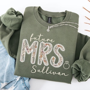 May include: An olive green sweatshirt with the words "future MRS Sullivan" in white script. The letters "MRS" are filled with a floral design. A silver necklace and a diamond ring graphic are also visible.