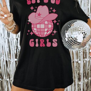 K&ouml;nnte beinhalten: Schwarzes T-Shirt mit einem pinkfarbenen Motiv einer Discokugel, die einen pinkfarbenen Cowboyhut tr&auml;gt. Der Text "Let's Go Girls" ist in Pink &uuml;ber dem Motiv geschrieben.