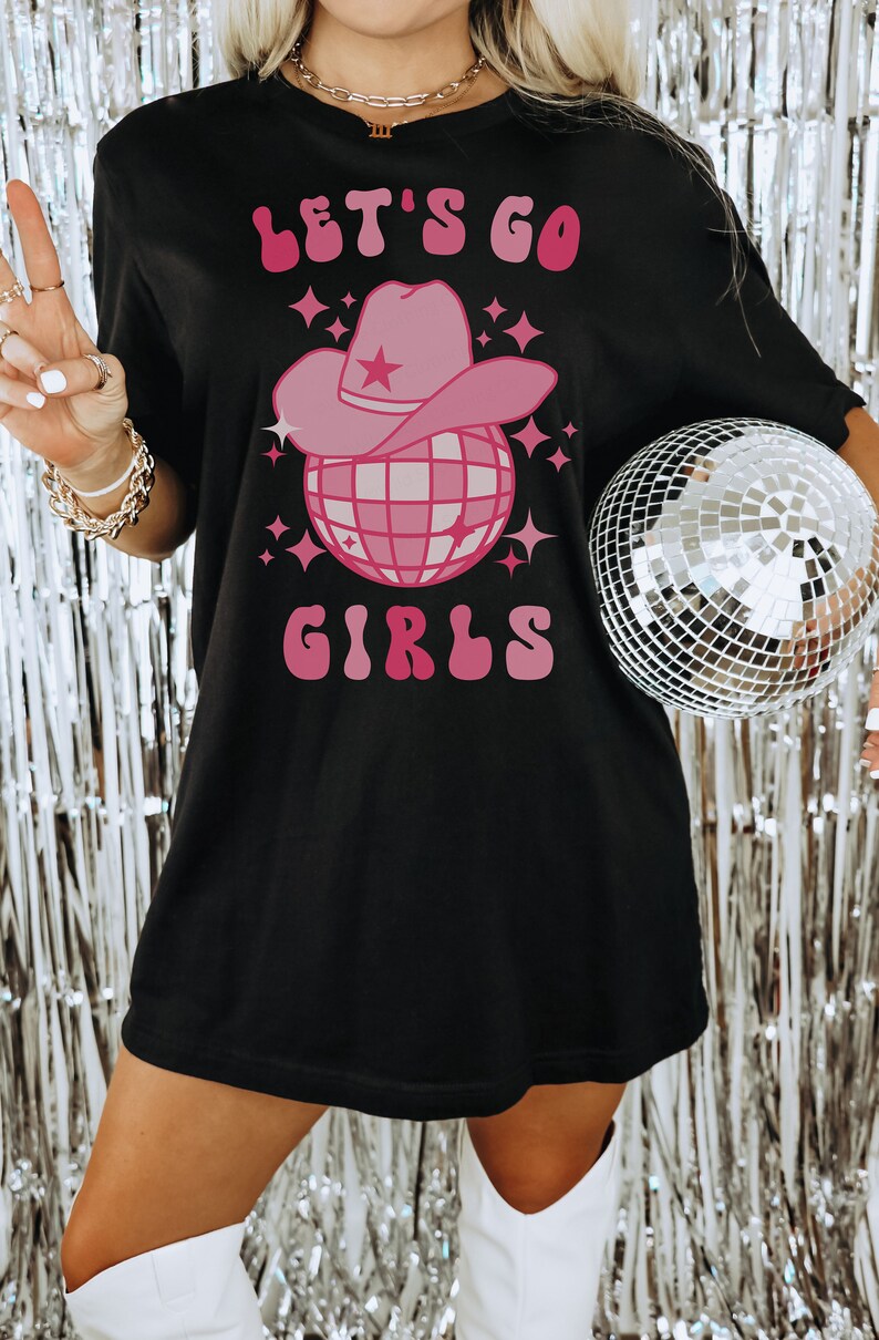 K&ouml;nnte beinhalten: Schwarzes T-Shirt mit einem pinkfarbenen Motiv einer Discokugel, die einen pinkfarbenen Cowboyhut tr&auml;gt. Der Text "Let's Go Girls" ist in Pink &uuml;ber dem Motiv geschrieben.