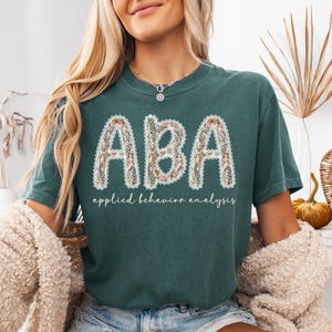 May include: Teal-colored t-shirt with the letters "ABA" in a floral design, outlined with white lace. Below the letters, the words "applied behavior analysis" are printed in a script font. The shirt is a casual style.