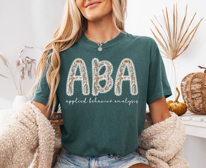 May include: Teal-colored t-shirt with the letters "ABA" in a floral design, outlined with white lace. Below the letters, the words "applied behavior analysis" are printed in a script font. The shirt is a casual style.