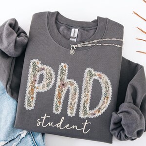 May include: A charcoal grey sweatshirt featuring the text "PhD student". The letters are decorated with a floral pattern and a white lace trim. A silver necklace with a pendant is draped across the top of the sweatshirt.