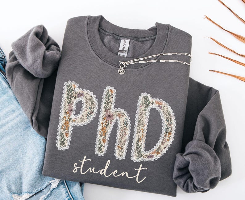 May include: A charcoal grey sweatshirt featuring the text "PhD student". The letters are decorated with a floral pattern and a white lace trim. A silver necklace with a pendant is draped across the top of the sweatshirt.