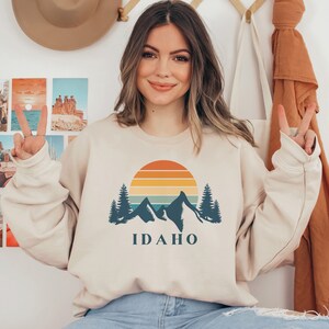 May include: A beige sweatshirt with a retro sunset graphic and mountains. The graphic includes a blue silhouette of mountains, trees, and a sun with orange, yellow, and red stripes. The text "IDAHO" is printed in blue below the graphic.
