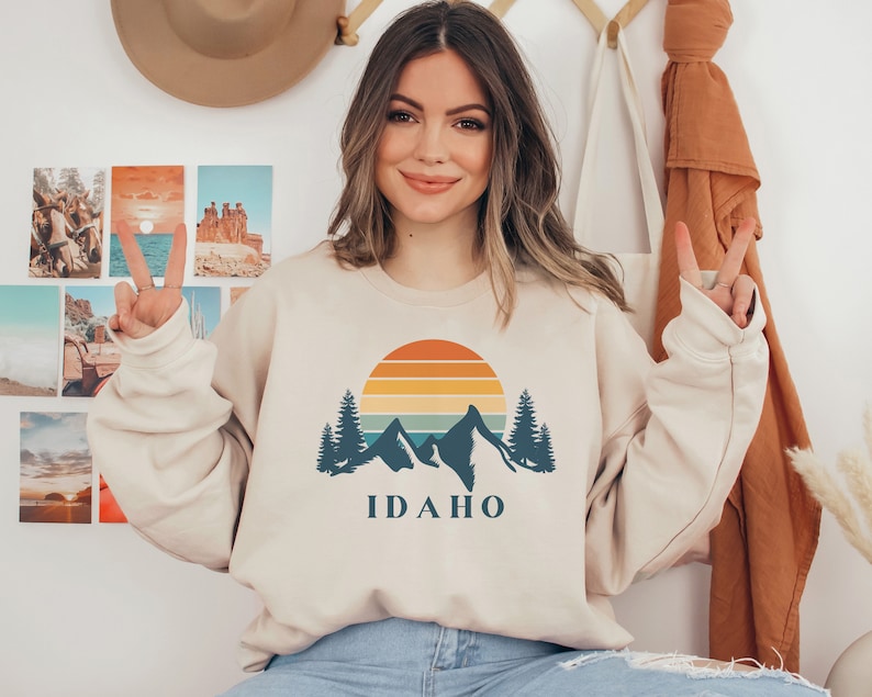 May include: A beige sweatshirt with a retro sunset graphic and mountains. The graphic includes a blue silhouette of mountains, trees, and a sun with orange, yellow, and red stripes. The text "IDAHO" is printed in blue below the graphic.