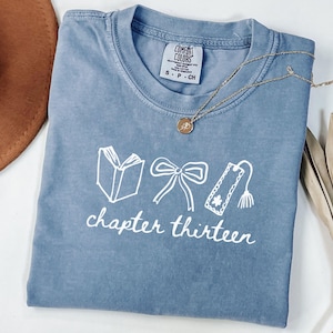 May include: A blue short-sleeved t-shirt featuring a white graphic of an open book, a bow, and a bookmark with a tassel. Below the graphics, the text reads chapter thirteen in white script. The shirt has a Comfort Colors brand label.