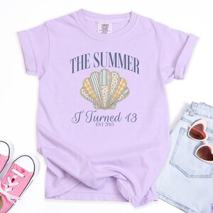 May include: A light purple short-sleeve t-shirt featuring a graphic of a seashell with various patterned sections. The text reads THE SUMMER I Turned 13 EST 2013. The shirt is displayed with pink canvas sneakers and light blue denim shorts with heart-shaped sunglasses.