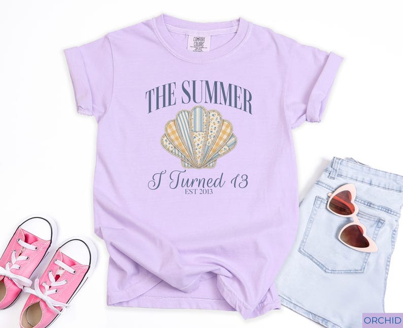 May include: A light purple short-sleeve t-shirt featuring a graphic of a seashell with various patterned sections. The text reads THE SUMMER I Turned 13 EST 2013. The shirt is displayed with pink canvas sneakers and light blue denim shorts with heart-shaped sunglasses.