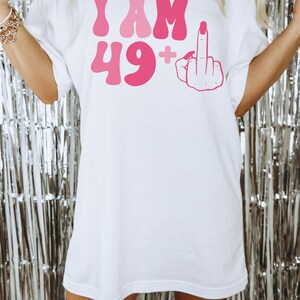 May include: White oversized t-shirt with pink lettering that says "I AM 49+" and a pink middle finger graphic.