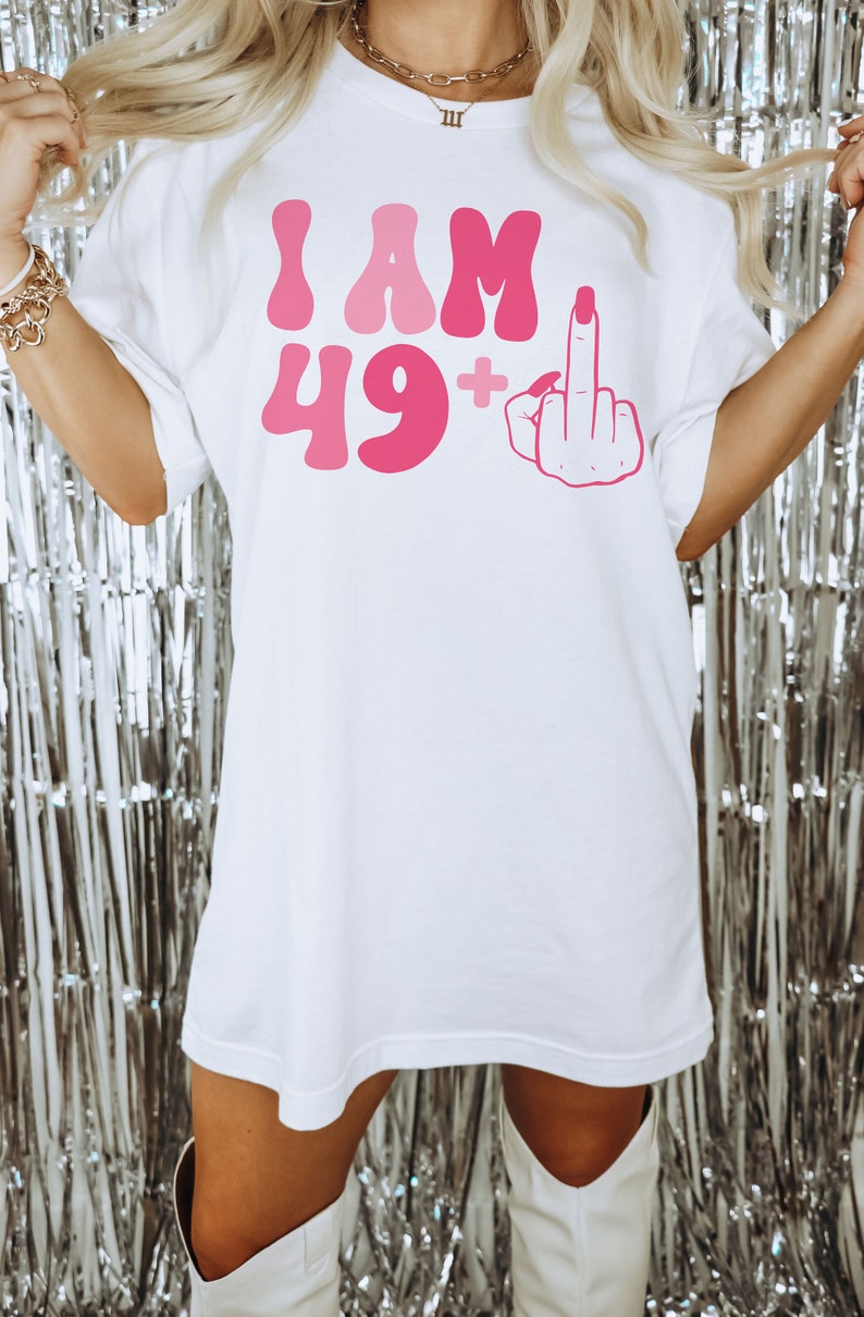 May include: White oversized t-shirt with pink lettering that says "I AM 49+" and a pink middle finger graphic.