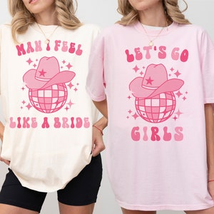 K&ouml;nnte beinhalten: Zwei T-Shirts mit rosa und wei&szlig;en Designs. Das erste Shirt ist elfenbeinfarben und sagt "Man I feel like a bride" mit einer rosa Discokugel mit einem rosa Cowboyhut darauf. Das zweite Shirt ist rosa und sagt "Let's go girls" mit einer rosa Discokugel mit einem rosa Cowboyhut darauf.