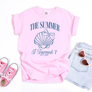 K&ouml;nnte beinhalten: Hellrosa T-Shirt mit dem Text "THE SUMMER I TURNED 7 EST 2019" und einer Muschel-Grafik. Dazu hellblaue Jeansshorts, rosa Sneakers und herzf&ouml;rmige Sonnenbrillen.