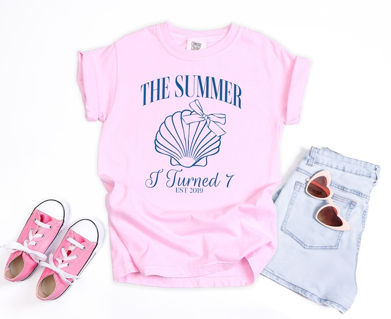 K&ouml;nnte beinhalten: Hellrosa T-Shirt mit dem Text "THE SUMMER I TURNED 7 EST 2019" und einer Muschel-Grafik. Dazu hellblaue Jeansshorts, rosa Sneakers und herzf&ouml;rmige Sonnenbrillen.