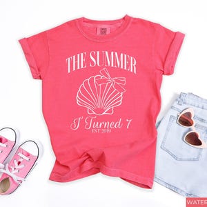 K&ouml;nnte beinhalten: Korallfarbenes T-Shirt mit dem Text "THE SUMMER I TURNED 7 EST 2019" und einer Muschel-Grafik. Das Shirt wird mit hellblauen Jeansshorts, einer rosa Sonnenbrille und rosa Turnschuhen kombiniert.