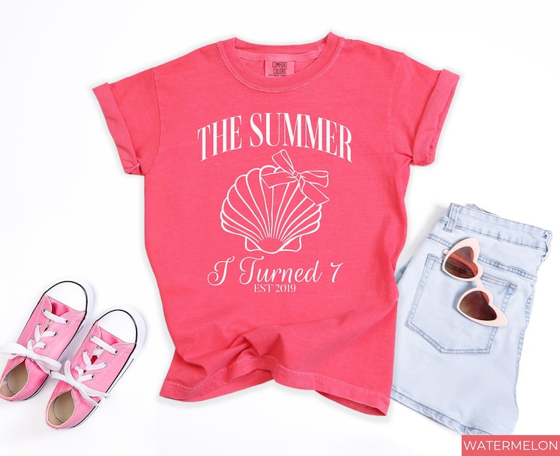 K&ouml;nnte beinhalten: Korallfarbenes T-Shirt mit dem Text "THE SUMMER I TURNED 7 EST 2019" und einer Muschel-Grafik. Das Shirt wird mit hellblauen Jeansshorts, einer rosa Sonnenbrille und rosa Turnschuhen kombiniert.