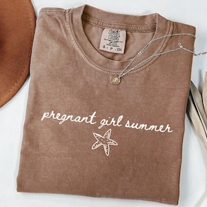 May include: A brown t-shirt with the text "pregnant girl summer" and a white starfish graphic. The shirt is made of a soft, comfortable material.