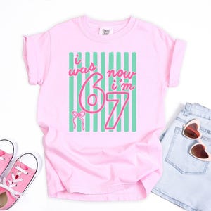 May include: A light pink t-shirt featuring a graphic with vertical teal stripes and the pink text "i was 6 now i'm 7" alongside a small pink bow illustration. The shirt has short sleeves, a crew neckline, and a tag at the inner collar area.