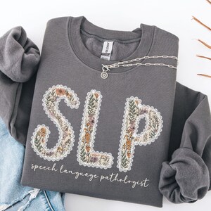 May include: A gray sweatshirt with the letters "SLP" in a floral design, with the words "speech language pathologist" below. A silver necklace with a pendant is draped across the sweatshirt.