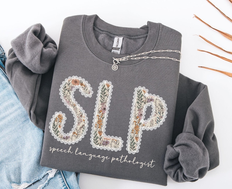 May include: A gray sweatshirt with the letters "SLP" in a floral design, with the words "speech language pathologist" below. A silver necklace with a pendant is draped across the sweatshirt.