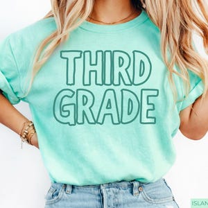 Pu&ograve; includere: Una t-shirt a maniche corte color verde menta con la scritta "THIRD GRADE" stampata in un carattere a contorno verde scuro sul petto. La maglia presenta un girocollo e una vestibilit&agrave; rilassata, con un design semplice e caratteri ben leggibili.