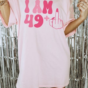 May include: Pink oversized t-shirt with the text "I AM 49+" and a pink cartoon hand giving the middle finger.