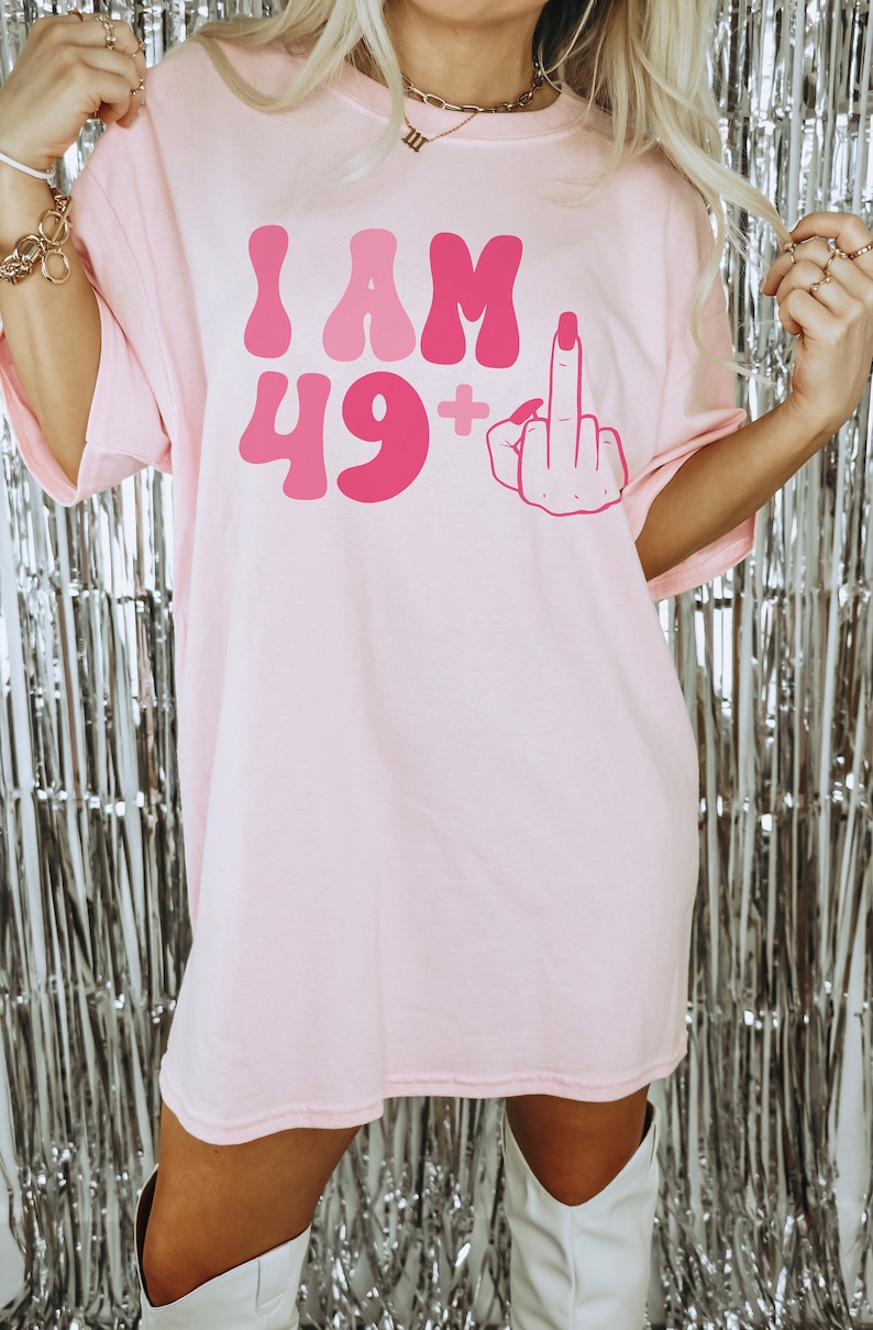 May include: Pink oversized t-shirt with the text "I AM 49+" and a pink cartoon hand giving the middle finger.