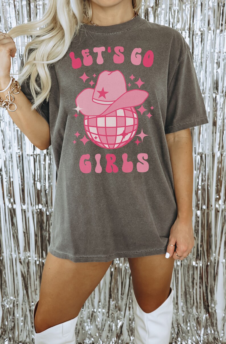 May include: A gray t-shirt with a pink graphic of a disco ball wearing a pink cowboy hat. The text on the shirt reads 'Let's Go Girls'.
