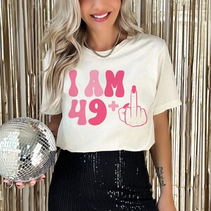 May include: White cropped t-shirt with pink lettering that says "I AM 49+" and a pink middle finger graphic.