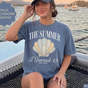 May include: A blue jean colored t-shirt featuring a central graphic of a seashell with a faux patchwork pattern in yellow, white, and blue. The text reads THE SUMMER I Turned 13 EST 2013 in white lettering. The shirt has a crew neckline and short sleeves.