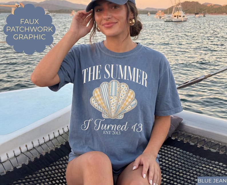 May include: A blue jean colored t-shirt featuring a central graphic of a seashell with a faux patchwork pattern in yellow, white, and blue. The text reads THE SUMMER I Turned 13 EST 2013 in white lettering. The shirt has a crew neckline and short sleeves.
