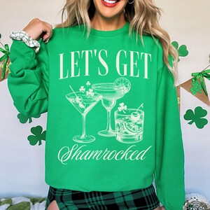 以下が含まれることがあります： 白い文字で「Let's Get Shamrocked」と書かれた緑色のスウェットシャツ。スウェットシャツには、クローバーのアクセントがついた3つのカクテルのグラフィックが描かれています。