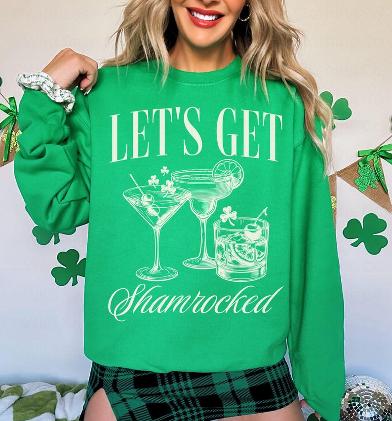 以下が含まれることがあります： 白い文字で「Let's Get Shamrocked」と書かれた緑色のスウェットシャツ。スウェットシャツには、クローバーのアクセントがついた3つのカクテルのグラフィックが描かれています。