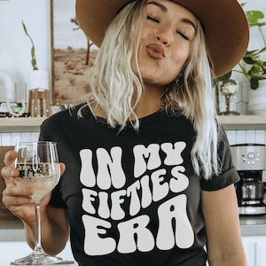 May include: A woman wearing a black t-shirt with white text that reads "In My Fifties Era" in a retro font. She is wearing a brown hat and has blonde hair. She is holding a glass of white wine.