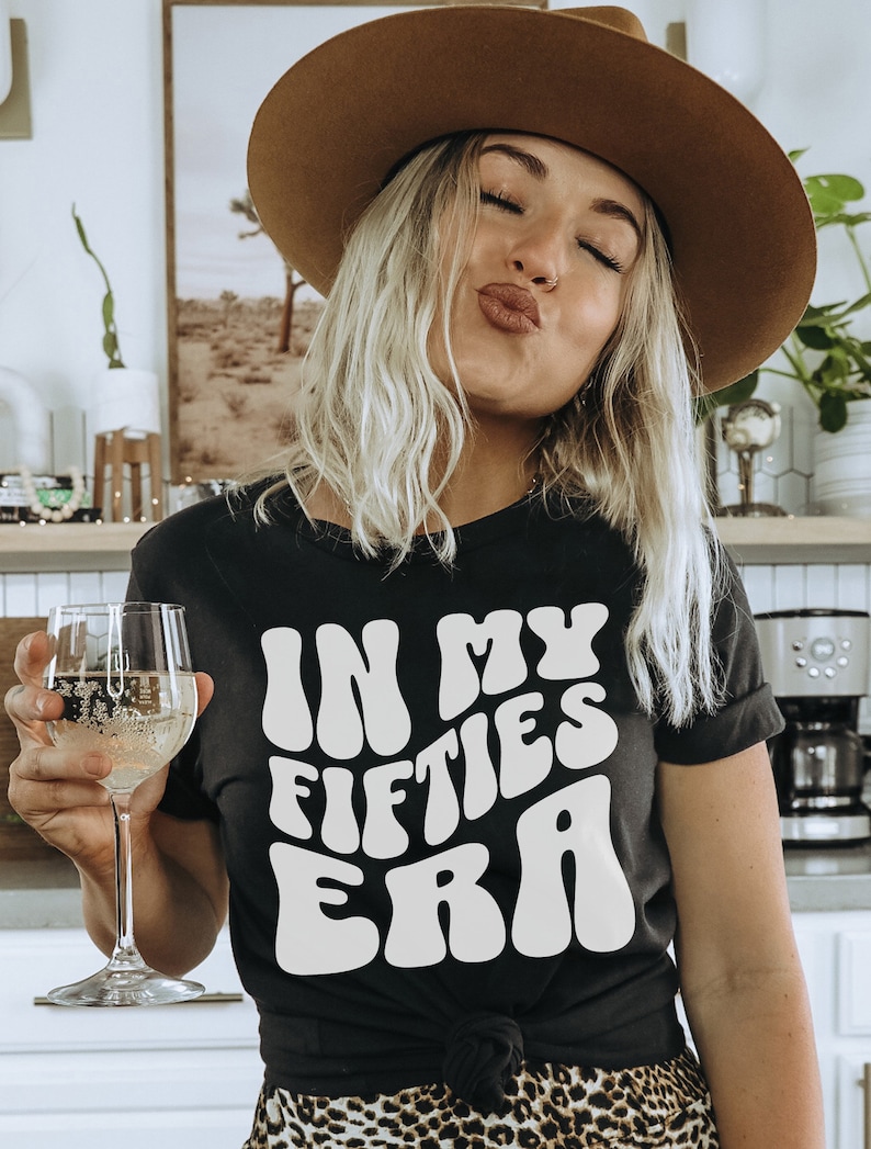 May include: A woman wearing a black t-shirt with white text that reads "In My Fifties Era" in a retro font. She is wearing a brown hat and has blonde hair. She is holding a glass of white wine.