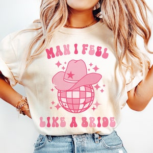 K&ouml;nnte beinhalten: Ein wei&szlig;es T-Shirt mit einem pinkfarbenen Grafikdesign, auf dem "Man I feel like a bride" steht, mit einer pinkfarbenen Discokugel und einem pinkfarbenen Cowboyhut darauf.