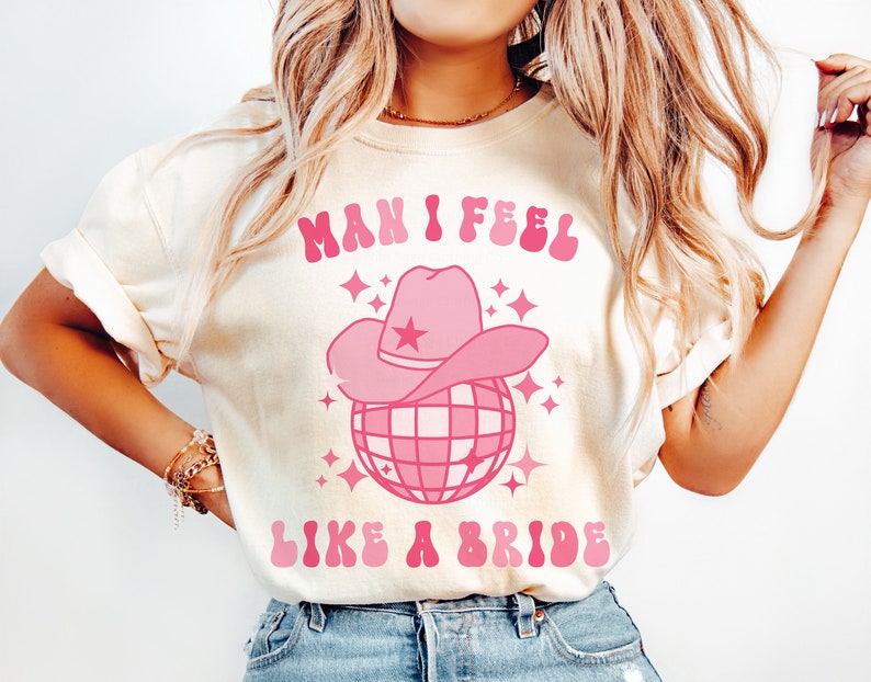 K&ouml;nnte beinhalten: Ein wei&szlig;es T-Shirt mit einem pinkfarbenen Grafikdesign, auf dem "Man I feel like a bride" steht, mit einer pinkfarbenen Discokugel und einem pinkfarbenen Cowboyhut darauf.