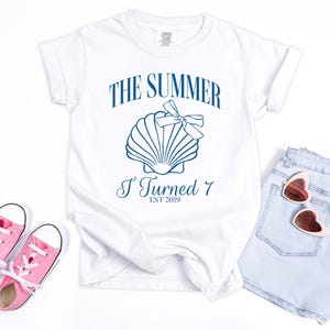 K&ouml;nnte beinhalten: Wei&szlig;es T-Shirt mit blauem Text "THE SUMMER I TURNED 7 EST 2019" und einer Muschel-Grafik. Rosa Turnschuhe und hellblaue Jeansshorts mit herzf&ouml;rmiger Sonnenbrille sind ebenfalls im Bild.