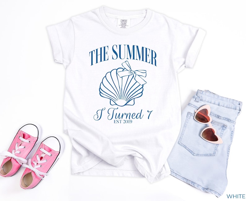 K&ouml;nnte beinhalten: Wei&szlig;es T-Shirt mit blauem Text "THE SUMMER I TURNED 7 EST 2019" und einer Muschel-Grafik. Rosa Turnschuhe und hellblaue Jeansshorts mit herzf&ouml;rmiger Sonnenbrille sind ebenfalls im Bild.