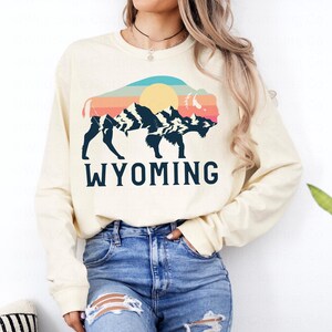 May include: A cream colored long-sleeved sweatshirt with a vintage-style graphic of a bison silhouette with mountains and a sun in the background. The text "WYOMING" is printed below the graphic.