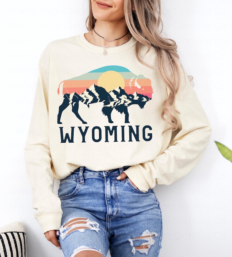 May include: A cream colored long-sleeved sweatshirt with a vintage-style graphic of a bison silhouette with mountains and a sun in the background. The text "WYOMING" is printed below the graphic.