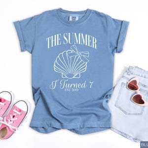 K&ouml;nnte beinhalten: Ein blaues T-Shirt mit dem Text "THE SUMMER I TURNED 7 EST 2019" und einer Muschel-Grafik. Das Shirt wird mit hellblauen Jeansshorts, einer rosa herzf&ouml;rmigen Sonnenbrille und rosa Turnschuhen kombiniert.